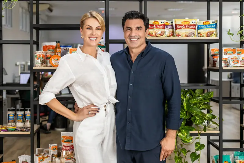 Ana Hickmann e Edu Guedes em pé, lado a lado, em frente a prateleiras de produtos