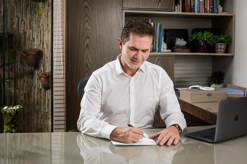 Cirurgião plástico de camisa branca assinando um documento à mesa do consultório